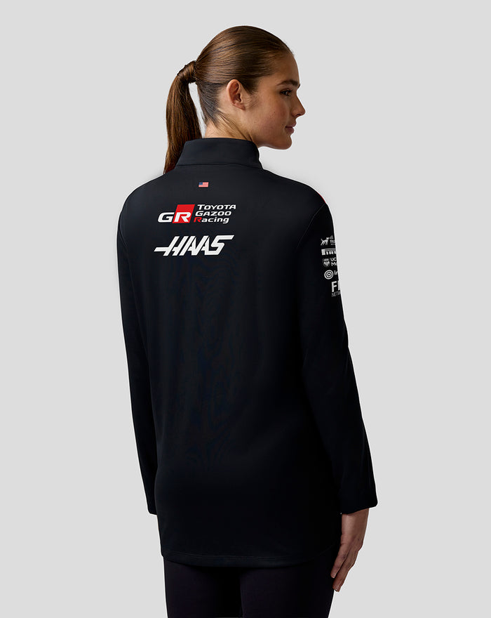 Back view of a model in a black Haas Team Quarter Zip Midlayer with sponsor logos on the back and a USA flag at the hem.