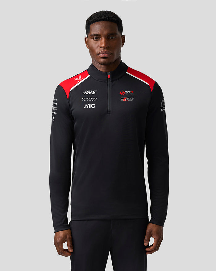 Front view of a male model in Haas Team Quarter Zip Midlayer, black with red shoulder panels and multiple sponsor logos on the chest.