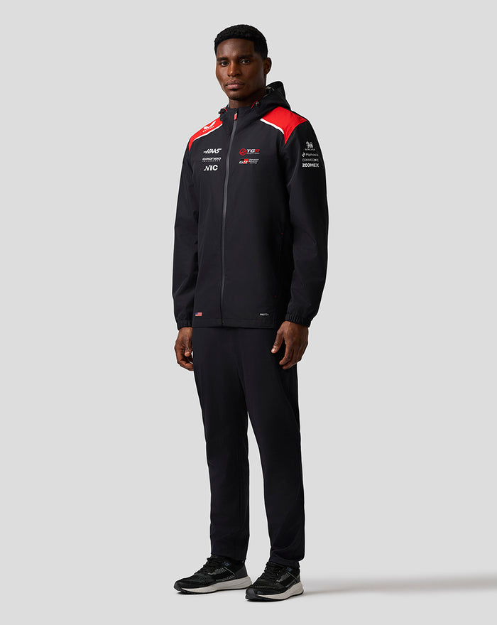 Model wearing Haas Team water-resistant jacket in black with red shoulder panels and hood, front zip and sponsor logos.