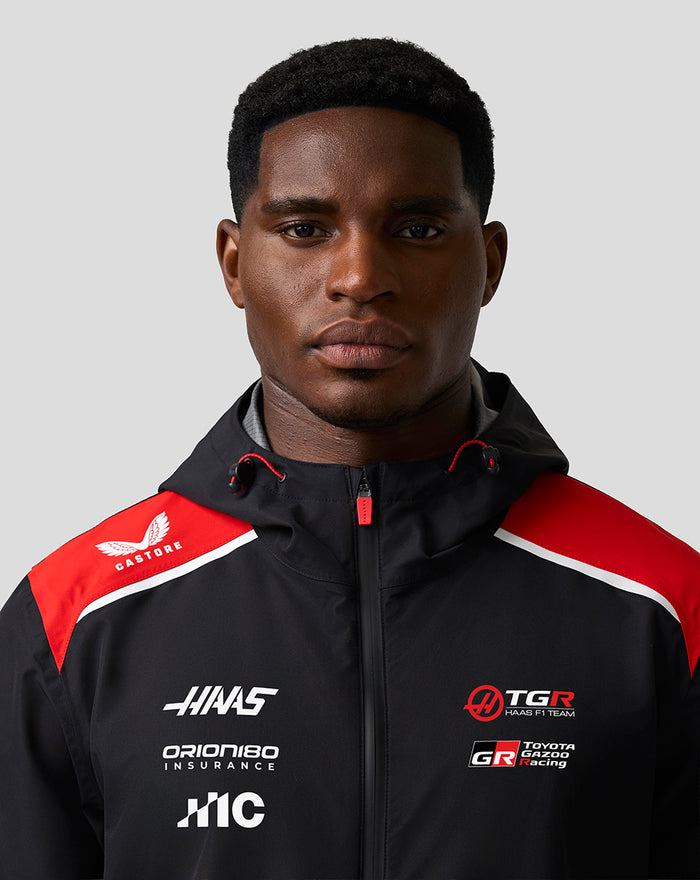 Male model wearing a black Haas Team water resistant jacket with red shoulder panels, hood, front zip and sponsor logos
