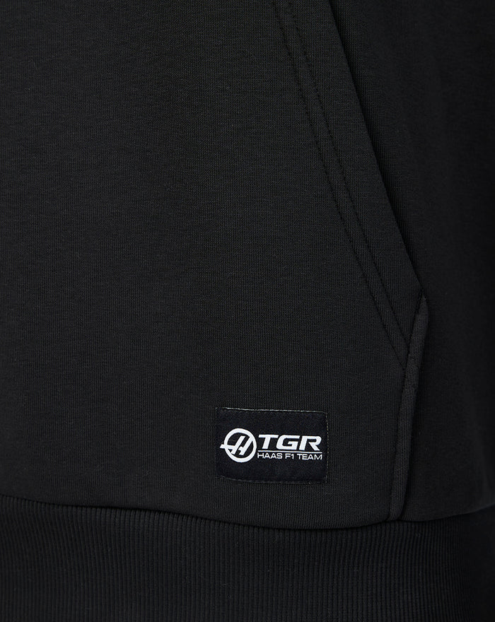 Close-up of black Haas hoodie fabric with a woven sponsor label at the lower left front hem.